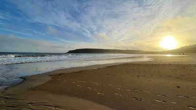 Sandy beach with footprints in the wet sand, white waves with the sun setting in the distance in the blue sky with some light clouds