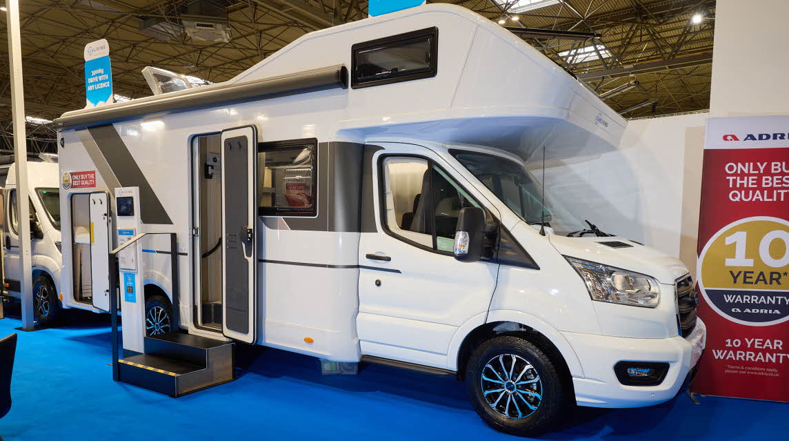 The Sun Living A Series 70DK is white with brown and bronze decals. There are two steps outside the open habitation door. There is an interactive stand next to the steps and the rear locker door is open.