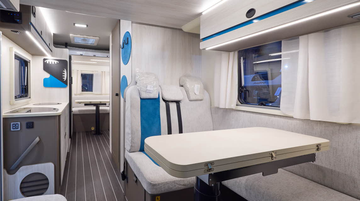 The Rimor Kilig 77 Plus has a wooden floor.  The upholstery is cream, with turquoise accents.  The cabinets are all very pale wood and the interior door is a dark brown.  There is a rear lounge and the vehicle has two separate tables which are shown 