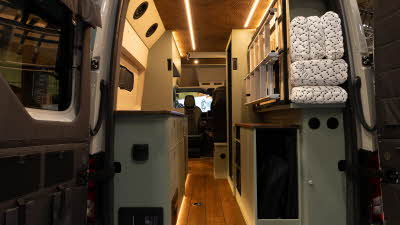 The Knights Custom Conversions Prestige XL has a wooden ceiling with a pattern, the cabinets are green with wooden edging.  The floor is wooden and there is a stowed ladder to the right.  The folded mattress is in the corner.