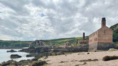 Porth Wen Brickworks Anglesey looking on from the sandy beach to the brick building on the right and the sea with seaweed covered rocks. The cliffs and green grass covered hills in the distance