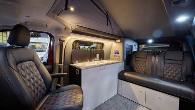 The Bespoke Campers Explore has a grey carpet, with pale grey cabinets.  The upholstery is black leather with red stitching.  Its pop top roof is fully extended. 