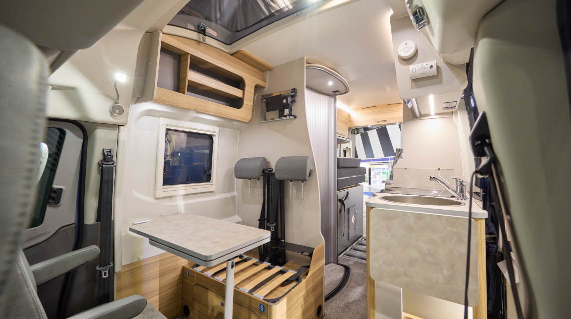 The Bailey Endeavour B65 has beige carpets, with wooden cabinets.  The upholstery is also grey.  The rear travel seats whose cushions are stored towards the rear reveal the storage area underneath.  The pop top roof is open.  