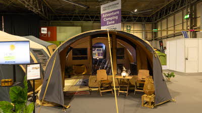 The Cabanon Mercury has brown canvas with yellow trim.  It has a curved roof design.  There is a wicker lamp with two chairs and a table in the awning.  The two internal bedroom windows are black.