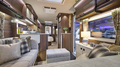 Interior of the Coachman LUSSO II with grey sofas, cosy cushions and wood accents and large windows.  There is a grey pouffe for extra relaxation. The door to the rear washroom is open. 