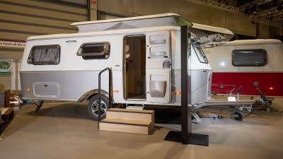 The classically styled Eriba Touring 630 Urban has a two tone layout in silver and white.  Its pop top roof is open as is its entrance door.  There are two wooden steps to gain easy access.