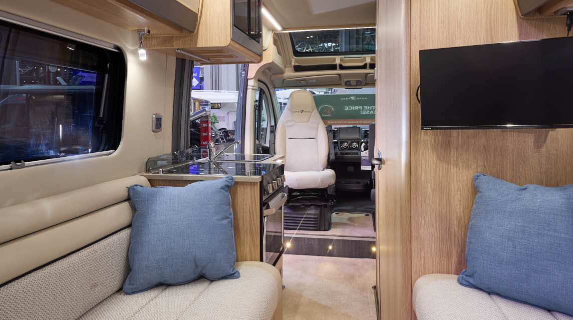 The Auto-Trail V-Line 540 SE has beige carpets, with pale wood cabinets with cream and grey overhead lockers.  The upholstery is white and cream.  There are two blue cushions.  The front seats are swivelled around.  There is a roof light.