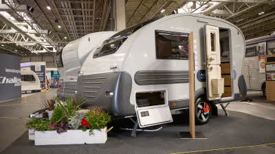 Adria Action exterior, white with dark grey bumpers and trim.  The locker door and main door are open and there are flowers around its hitch in white planters.   
