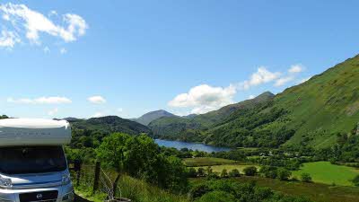 In the bottom left there is a motorhome parled beside a fence next to lush green fields and hills with  a lake in the middle and mountains in the distance
