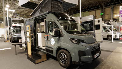 The Adria Twin Sports 600 SPB is grey, its rising roof is extended.  The sliding door is open and there are two steps to gain easy access.  There is an interactive stand.