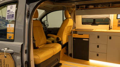The Wildtracks Campervans Panoramic has tan leather upholstery and has grey cabinets.  There is a black fridge.