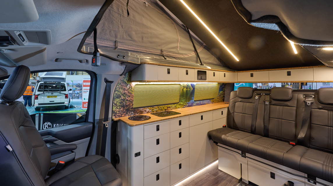 Knights Custom Conversions Prestige Tourer Plus has wooden flooring with black upholstery.  The cabinets are white and pale wood.  The pop top roof is open.