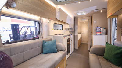 Adria Adora Seine’s interior, it has wooden cabinets with grey upholstery in the lounge.  There are three cushions, blue, green and purple.  The kitchen has a domestic style oven and hob.  The view leads to the bedroom with 3 skylights. 