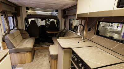 The Bailey Autograph 72-2 has beige upholstery with a brown leather surround.  The kitchen is to the right.  The drop down bed is in the up position above the lounge.  There is removable beige carpet.