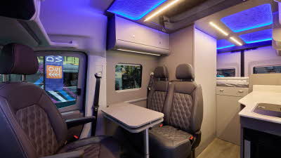 The Bespoke Campers Edition (Four Berth) has a wooden floor, with pale grey cabinets.  The upholstery is brown leather.  There is a fixed rear bed.  The pop top roof is open and it has blue lighting.