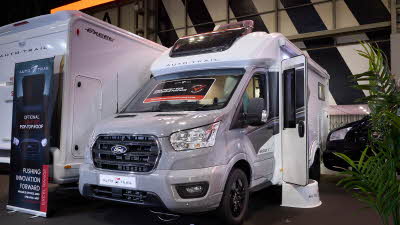 The Auto-Trail Excel 690T has a silver cab and white body with grey decals.  There is a show offer on its windscreen in red.  There is a white step by the opened habitation door.  It has a large sunroof and its roof light is open.