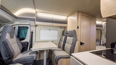 The Auto-Trail Expedition 67 has grey flooring, with pale wood cabinets with pale wood and grey overhead lockers.  The upholstery is grey and cream.  There is a rear lounge and the rear doors are open.