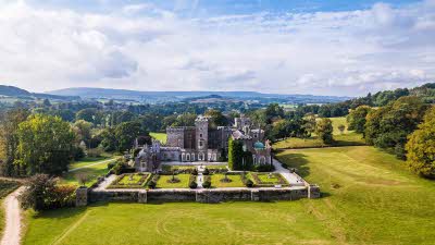 A view from above of Powderham Castle surrounded by the castle walls amongst the lush green fields with a view of the hills in the distance