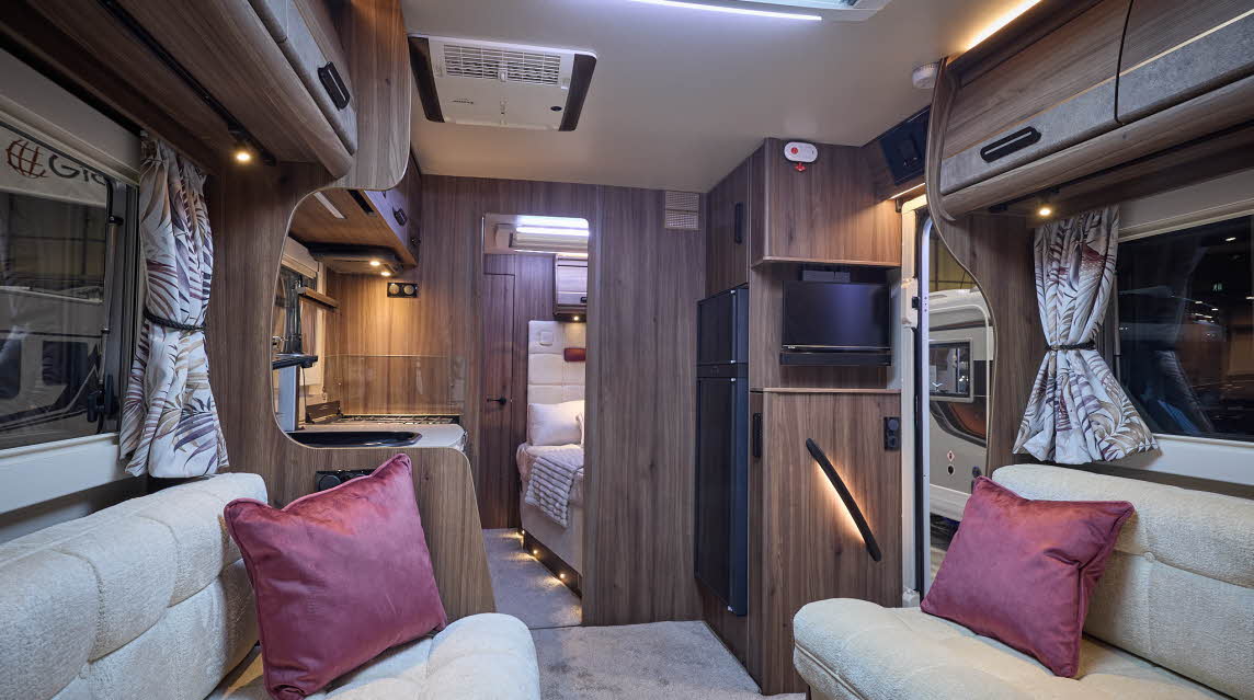 The Auto-Sleeper Burford has a pale grey carpet, with dark wood cabinets. There is a roof light. The upholstery is cream with two burgundy cushions. There is a fixed bed to the rear.