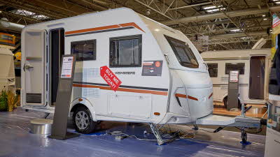 Weinsberg CaraOne 390 QD is white with orange and black decals.  Its entrance door is open and there is a metal step to gain easy access.  There is an information board next to the step.
