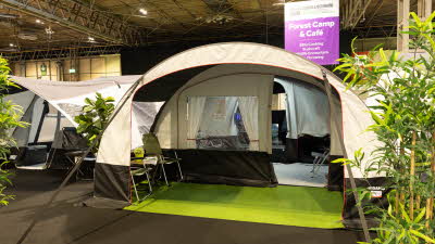 The Trigano Nebra has cream and brown canvas.  It has a curved structure.  There are two green chairs in the awning area which has a faux grass mat.  The entrance door to the living area is open.