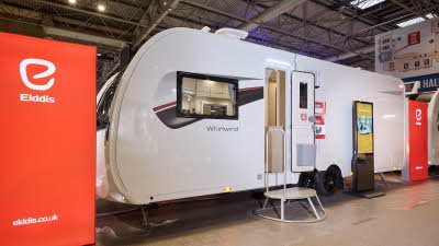 The Elddis Whirlwind 860 has a white exterior with a grey, red and black decal. The entrance door is open and there is a metal step to gain easy access. There is a large Elddis red banner with their website address next to it.