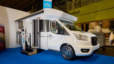 The Sun Living S Series 75SL is white with brown and bronze decals.  There are two steps outside the open habitation door.  There is an interactive stand next to the steps and the rear locker door is open.