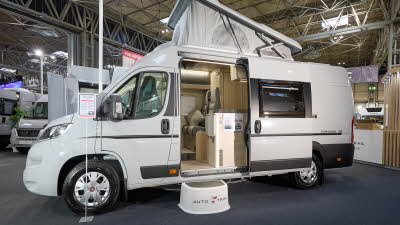 Auto-Trail Expedition 68's exterior is white with dark grey decals.  The sliding door is open revealing the lounge and kitchen.  There is a white step to gain easy access.  The rising roof is extended.