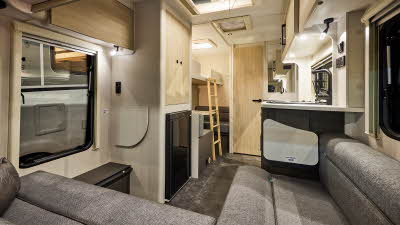 Interior of the Xplore XC LITE with grey sofas, wood accents and grey carpets.  The large windows and roof light will let natural light flood in.  There is a ladder to the single bunk situated above the dinette. 