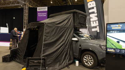 The TentBox Classic has black canvas and a black hard shell roof.  It has an annex and the door is open.  It is shown on a grey vehicle.  There are two storage boxes located outside the annex.