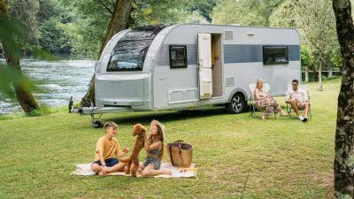 Adria Altea caravan on a grassy bank beside a river with a family sat on the grass 