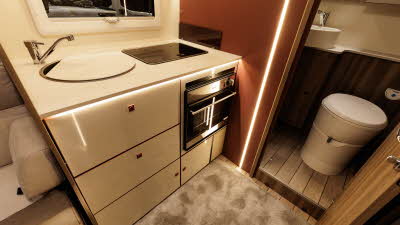 The Roller Team Kronos Compact 590 has a rear kitchen with an oven.  The round sink has chopping board cover.  The washroom is next to it, the door is open, revealing the fitted toilet.