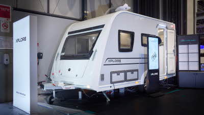 Xplore XC LITE's exterior is white with black/turquoise decals.  It has large windows.  The roof light is open.  There is an interactive display on the left