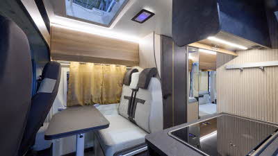 The Rimor Horus 40 has a wooden floor.  The upholstery is grey and dark brown.  The cabinets are all pale wood.  The skylight is open.  The covered hob and sink are at the front.  There is a rear lounge and the rear door is open.