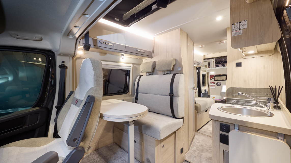 The Elddis Autoquest CV80 has beige carpets and cream and pale brown cabinets.  The upholstery is cream and pale brown.  The round sink is on the right.  There is a rear lounge with two sofas.  The rear doors are open.