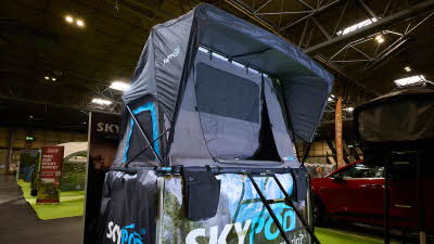 The SkyPod Rapide has black canvas walls and has turquoise flashes around it.  The interior canvas is blue.  There are two support rods by the window.  It is shown on a display plinth.  There is a black ladder.