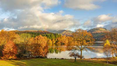 Autumn colour on the trees around Windermere in the Lake District, with a rainbow faintly visible in the clouds as they clear away