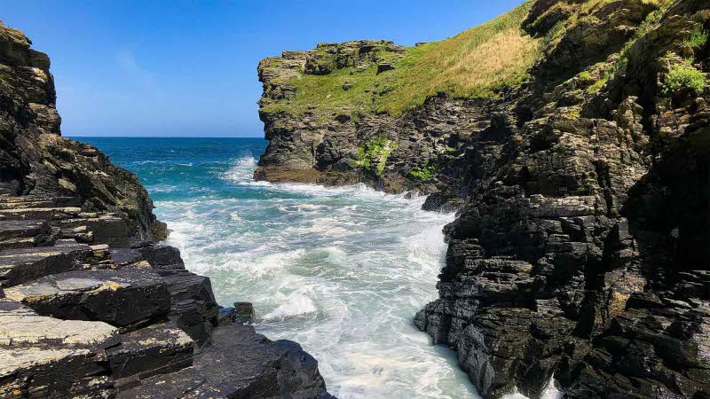 White water rushing in from the blue sea between rocky walls with a back drop of a clear blue sky meeting the sea. 