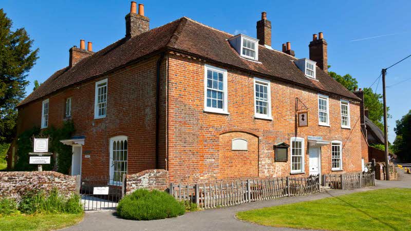 An up close view of Jane Austen's House Museum in Chawton. The red brick building with many windows and 5 chimney stacks. A pathway runs along the front of the house leading to green grass under the lovely blue sky