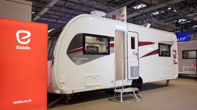 The Elddis Crusader Shamal has a white exterior with grey, red and black decals.  The entrance door is open and there is a metal step to gain easy access.  There is a large Elddis red banner with their website address next to it.