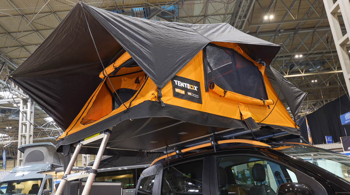 TentBox Lite 2.0 has orange canvas with a black canvas roof.  It has a platform.  It is shown on a vehicle with orange roof rails.  Its door is open.