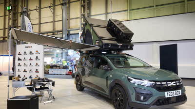 TentBox Cargo 2.0 is shown on a Dacia. The canvas is green and it has a black hard top with a storage box. There is an awaning and there are table and chairs underneath.