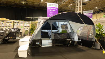 The Jamet Eco Compact has cream and brown canvas.  It has a curved structure.  There is a chair and table in the awning area.  The entrance door and large window in the living area are open.