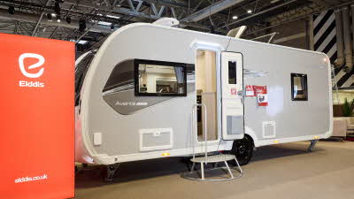 The Elddis Avanté APEX 550 has a grey exterior with a grey and black decal.  The entrance door is open and there is a metal step to gain easy access.  There is a large Elddis red banner with their website address next to it.