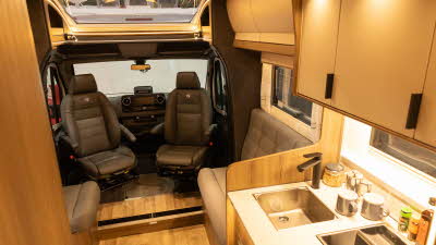 The Coachman Sportivo 565 has beige upholstery with wood furniture.  The locker doors are cream.  The kitchen is to the right and has a square sink.  There is a large sunroof above the cab.  