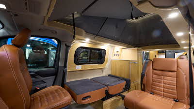 The Rolling Homes Camper Ltd Stanley has dark grey flooring. The upholstery is brown leather. The cabinets are wooden. The rising roof is fully extended. One of the passenger seats is folded down.