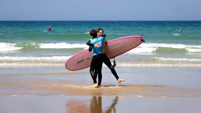Two surfers hugging with one holding a surfboard, standing in the shallow water with small waves, shallow green water leading to the deeper blue as the sky meets the sea