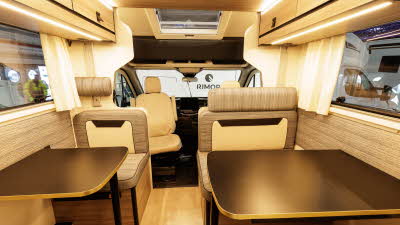 The Rimor Kilig 79 has cream/beige patterned upholstery.  There are two tables in front of the rear seats.  There is accent lighting throughout.