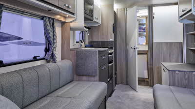 Swift Sprite Alpine 2’s interior has grey upholstery with wooden furniture.  The kitchen is in the middle and its furniture has dark doors. .  The rear door is open revealing the washroom