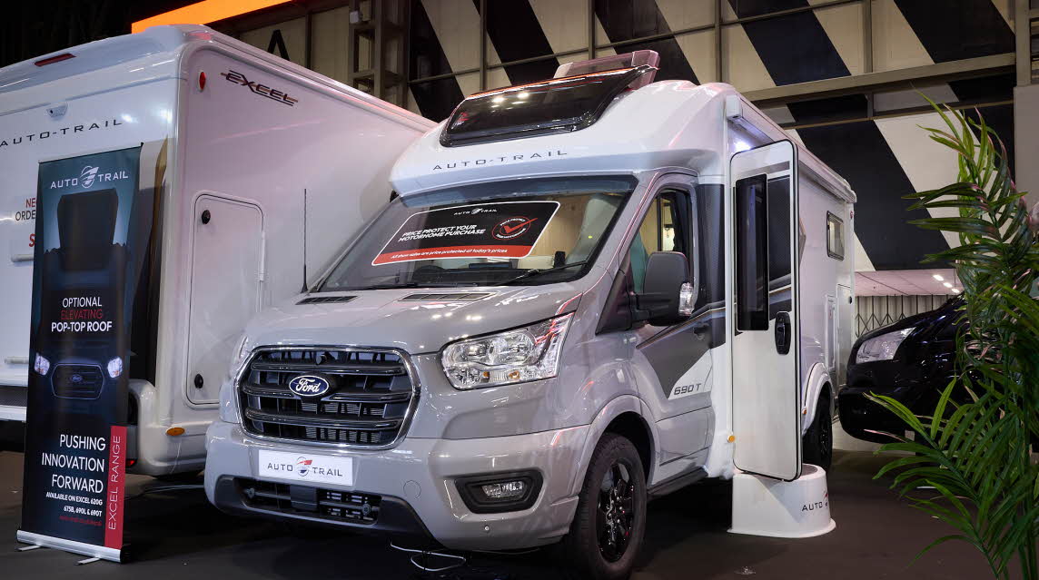 The Auto-Trail Excel 690T has a silver cab and white body with grey decals.  There is a show offer on its windscreen in red.  There is a white step by the opened habitation door.  It has a large sunroof and its roof light is open.